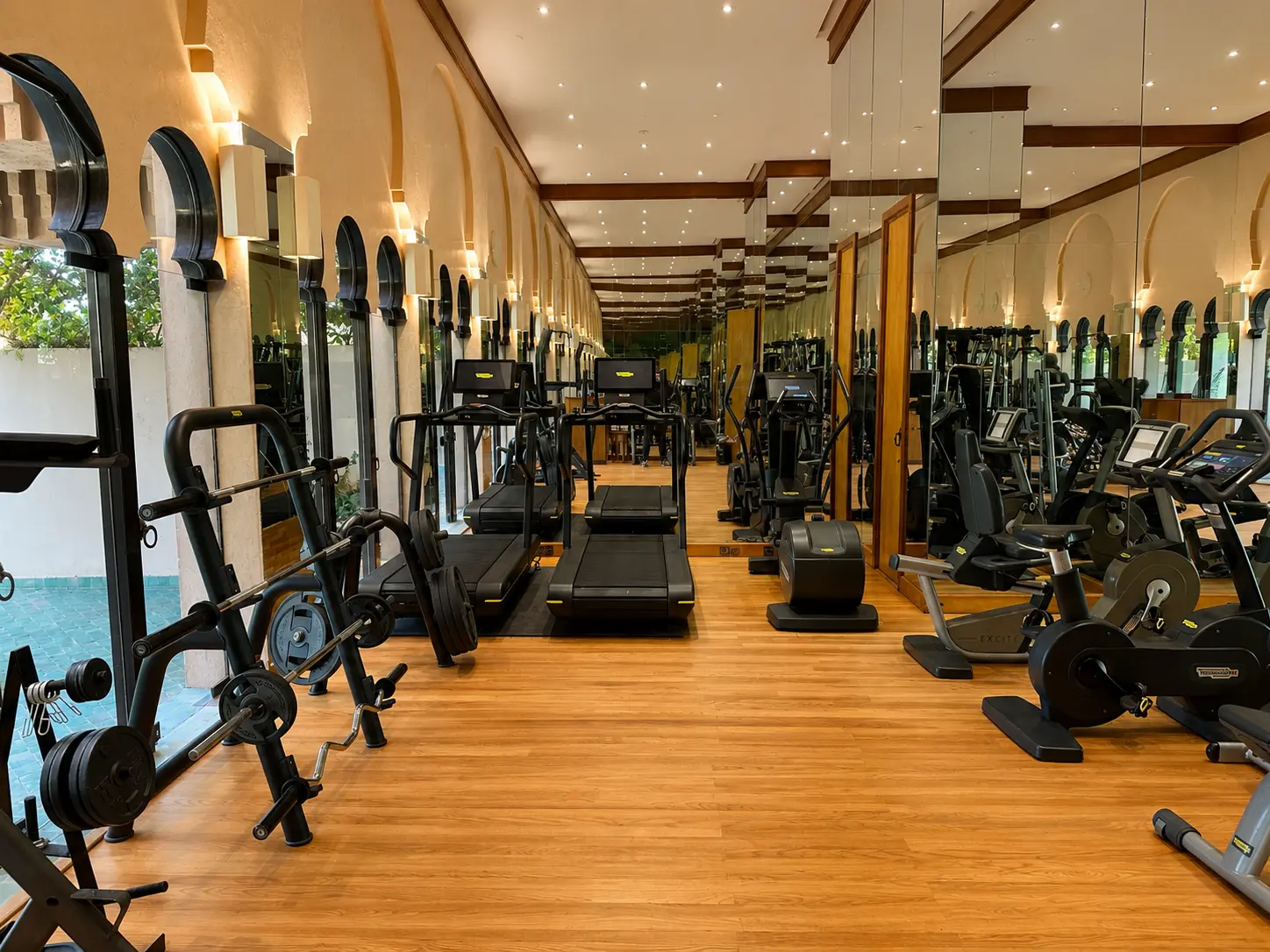 The gym at Amanjena in Marrakech with Technogym cardio machines, free weights, mirrored walls, wooden floors, and Moroccan arched windows.