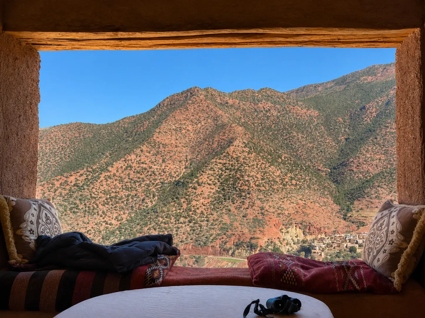 Panoramic view of the Atlas Mountains and a traditional Berber village in Morocco, framed by a rustic window seat overlooking the Ourika Valley.