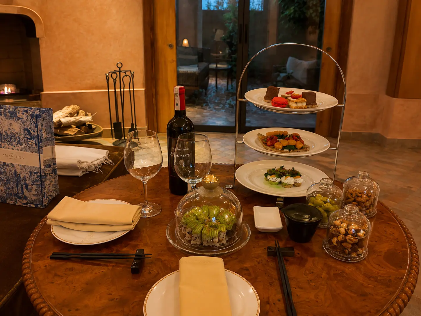 Private in-room dining setup at Amanjena in Marrakech with Japanese dishes, desserts, wine, snacks, and a fireplace inside a Pavilion.