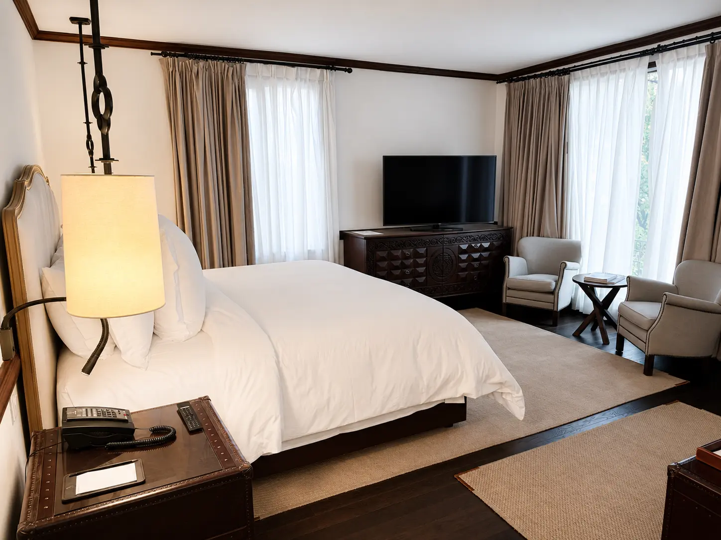 Luxury hotel bedroom with white linens, dark wood furniture, floor-to-ceiling windows and classic neutral interiors.