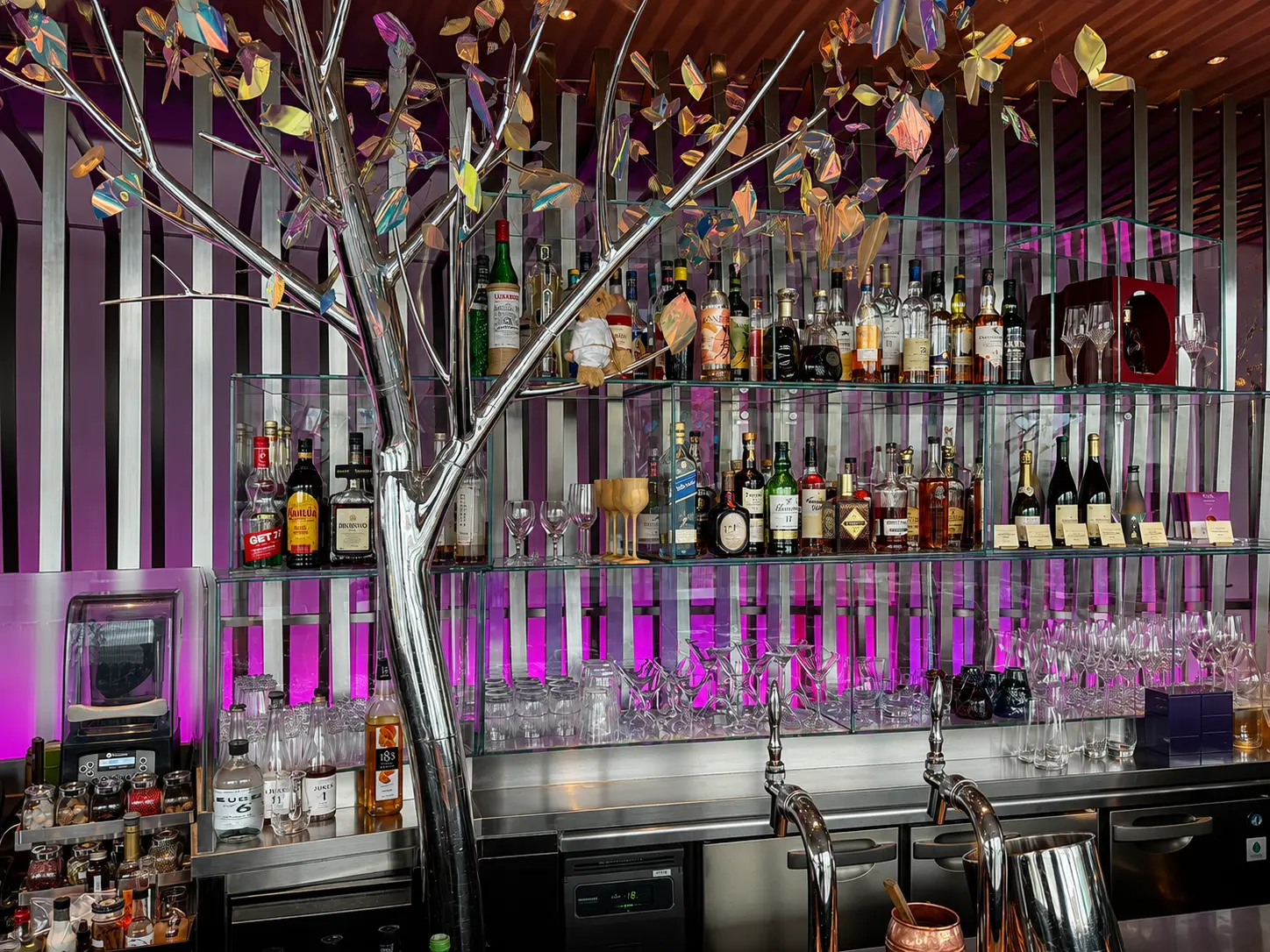 Interior of Peter: The Bar at The Peninsula Tokyo with a chrome tree sculpture, colorful leaves and backlit drinks display.