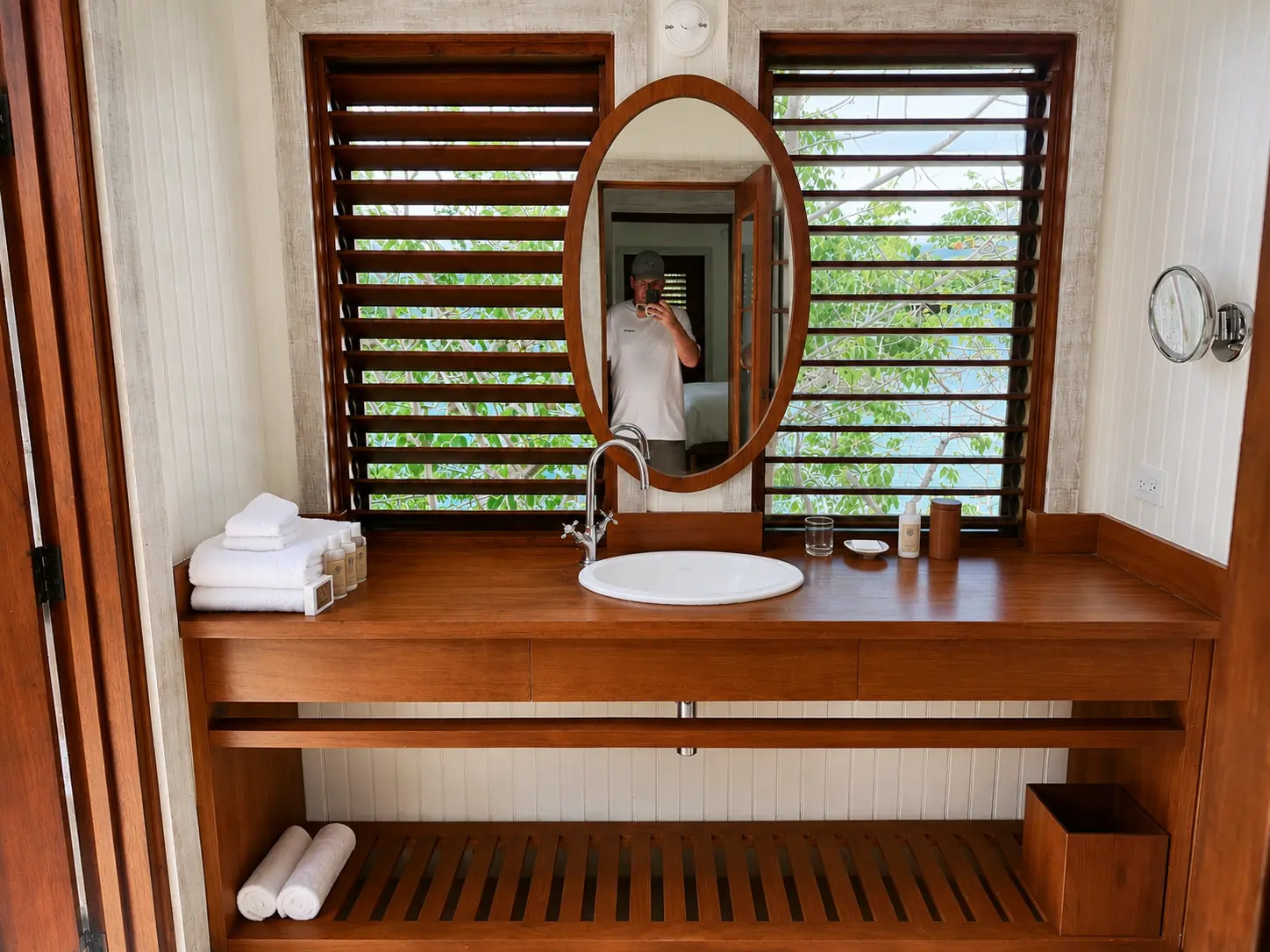 Bathroom vanity inside a One Bedroom Beach Hut at GoldenEye Jamaica with wooden counter, oval mirror and louvered windows.