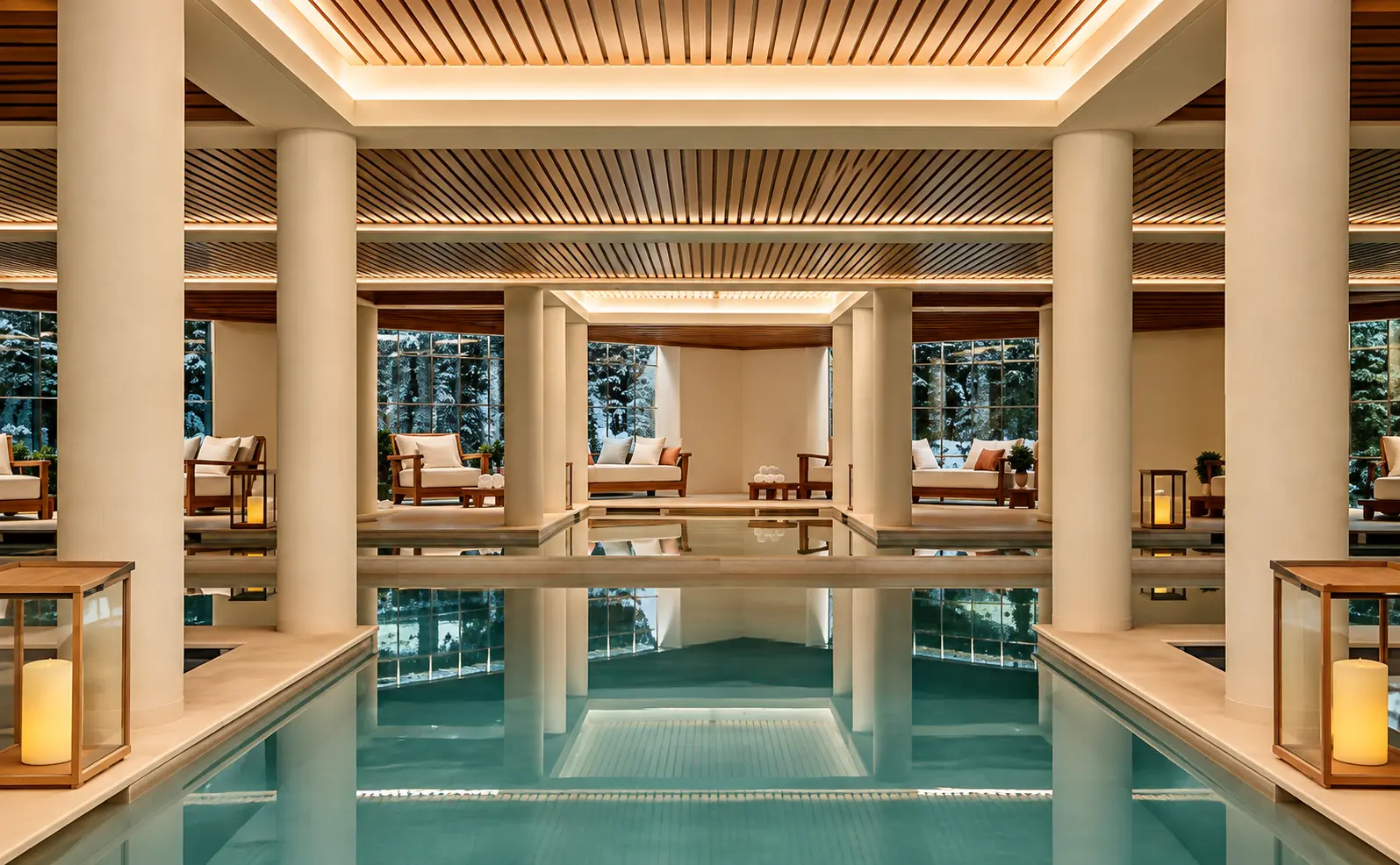 Indoor swimming pool and spa at Aman Le Mélézin with timber ceilings, cream columns and snowy forest views.