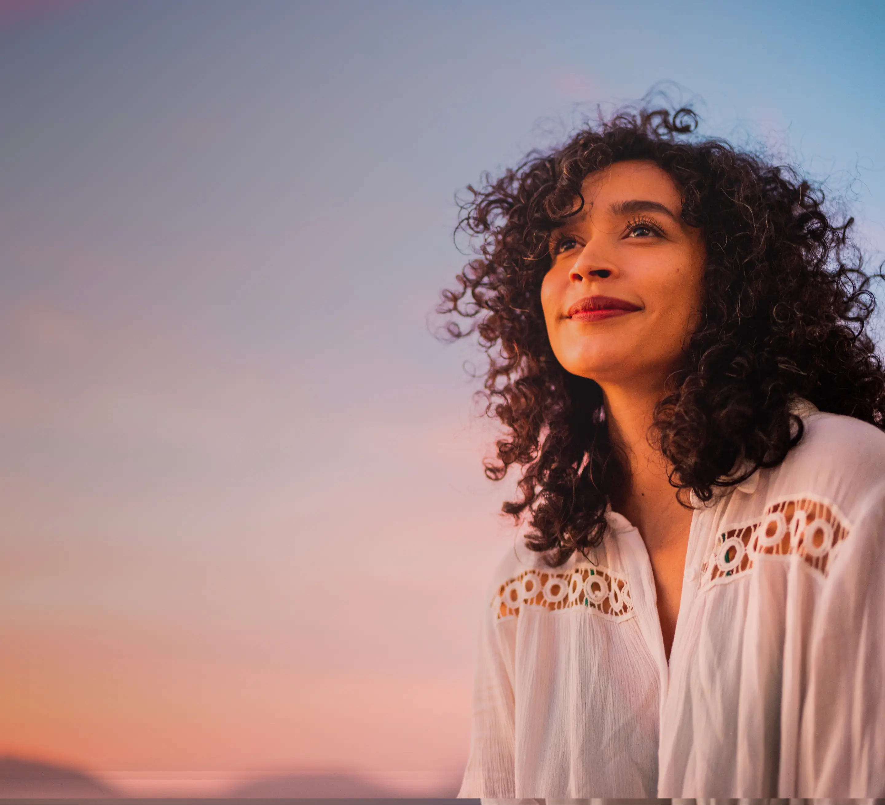 Woman with curly hair smiling and looking up at a soft pastel sky