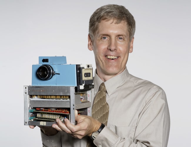 Man in beige shirt and tie holding an old blue camera with a cassette tape slot.