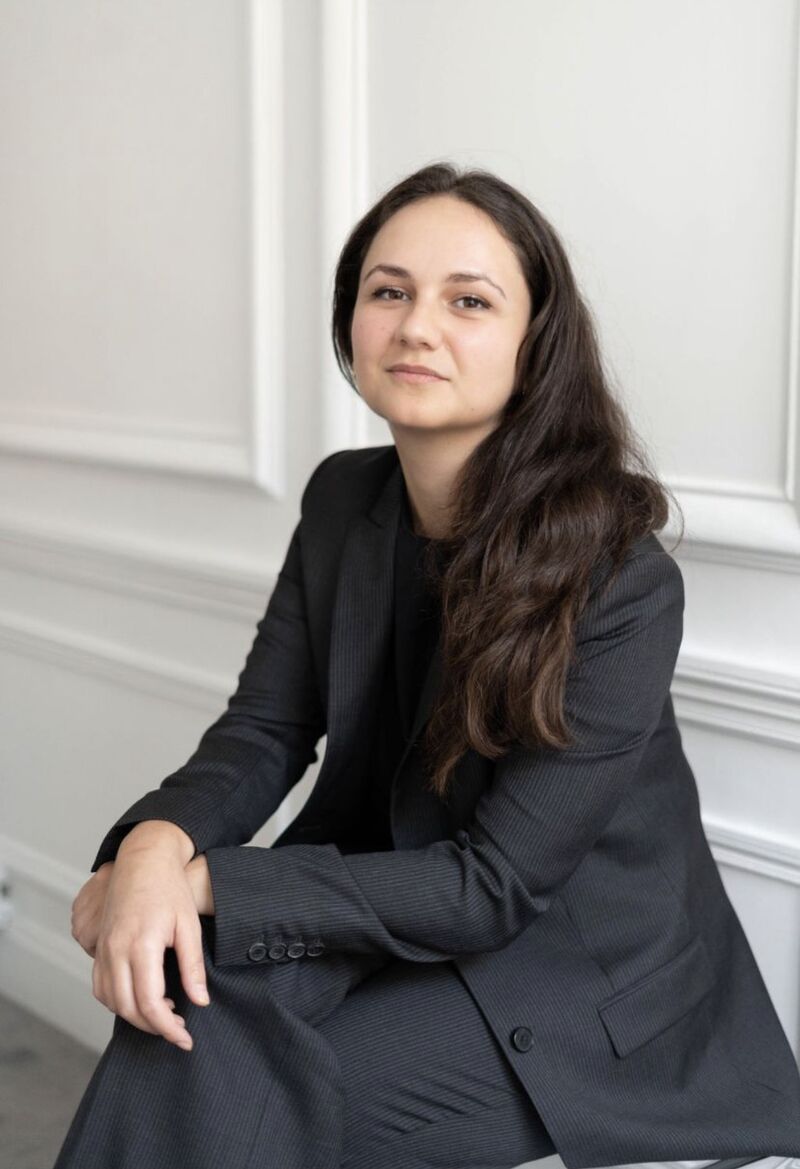 Jeune femme aux cheveux longs et bruns portant un costume gris foncé assise avec un léger sourire dans un intérieur clair.