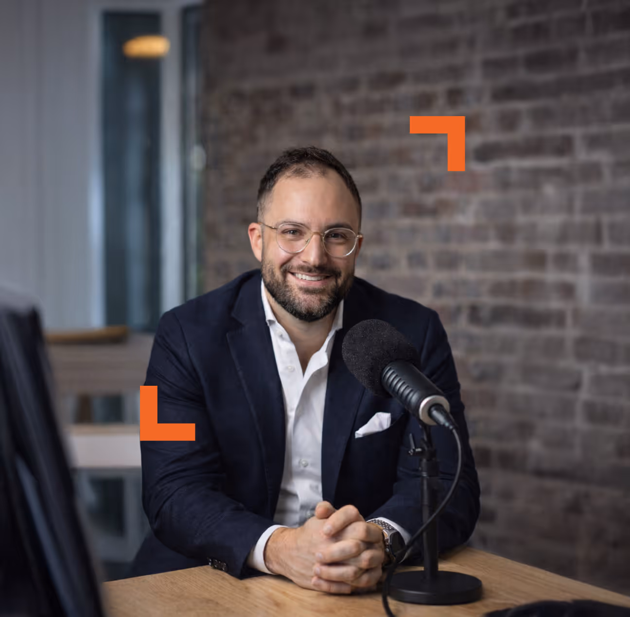 Smiling man with glasses and beard sitting at a table with a microphone in front of a brick wall.