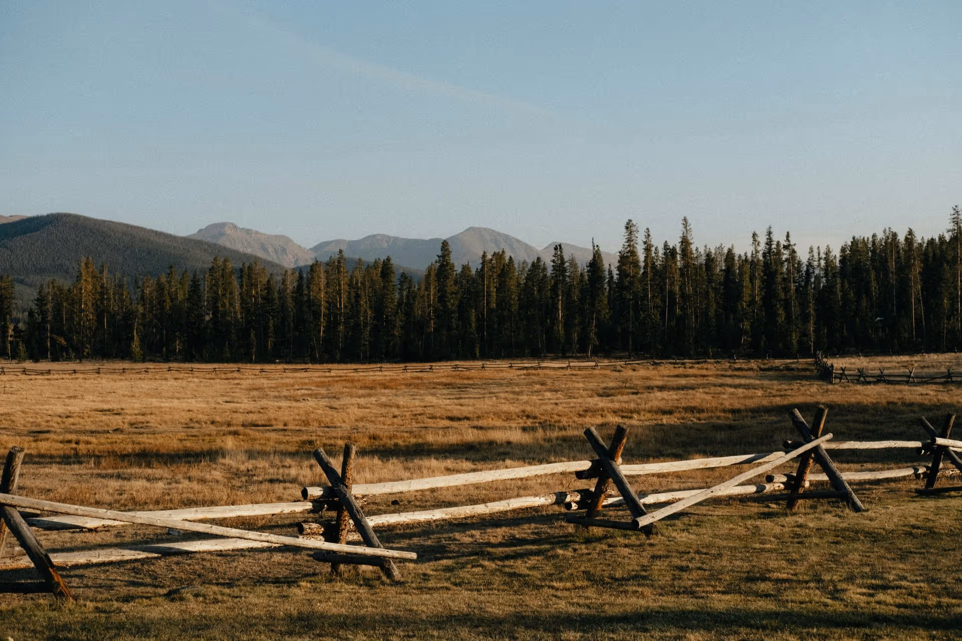 Wooden fence along a grassy field with forest and mountains in the background under a clear sky.