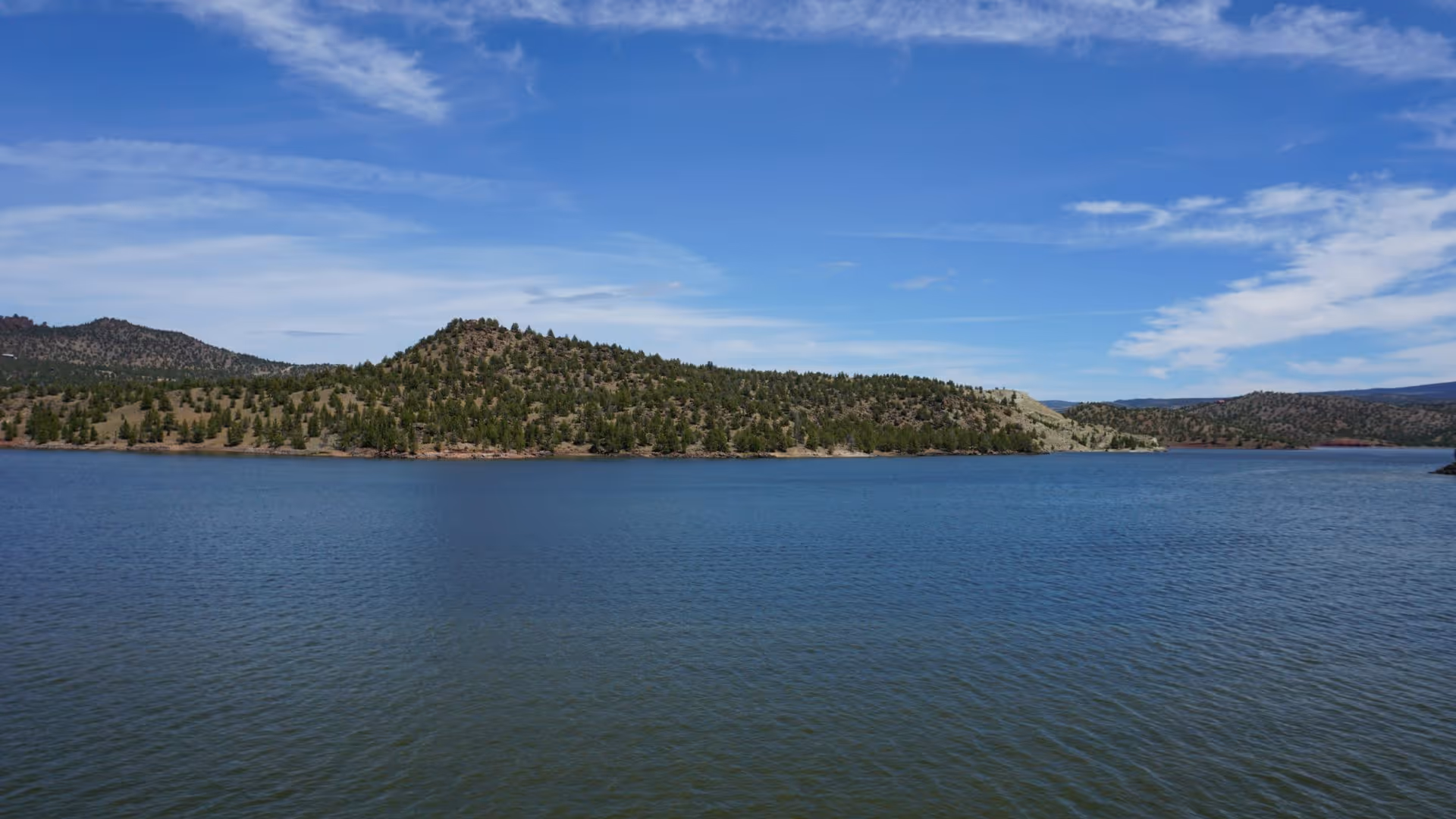 Lake with calm water surrounded by tree-covered hills under a partly cloudy blue sky.