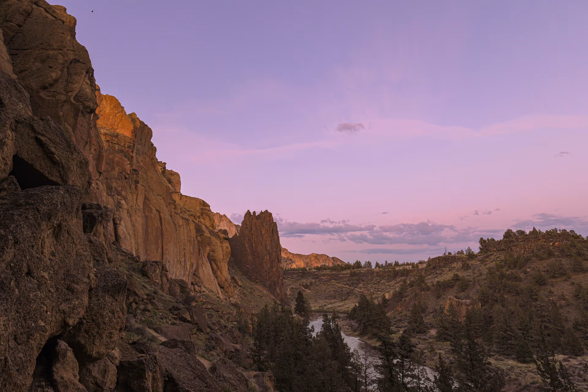 Rocky cliffs and pine trees along a riverbank under a purple sunset sky.