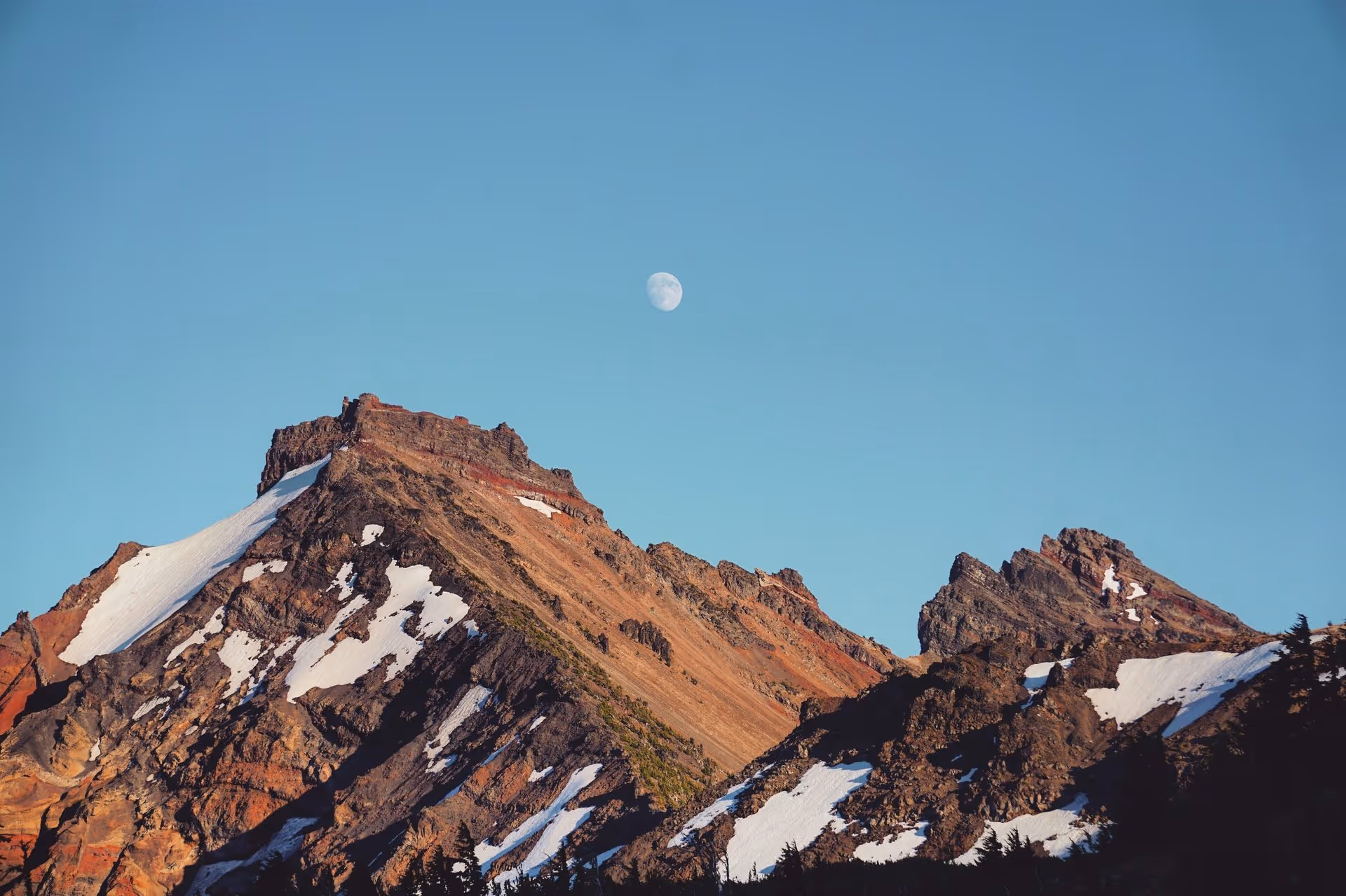 Rocky mountain peaks with patches of snow under a clear blue sky with a visible moon.