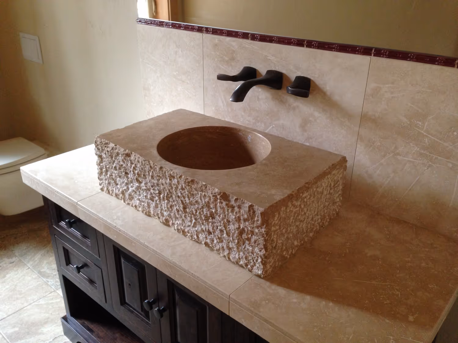 Brown stone sink with rough edges on a tiled countertop and dark wooden cabinet below in a bathroom.