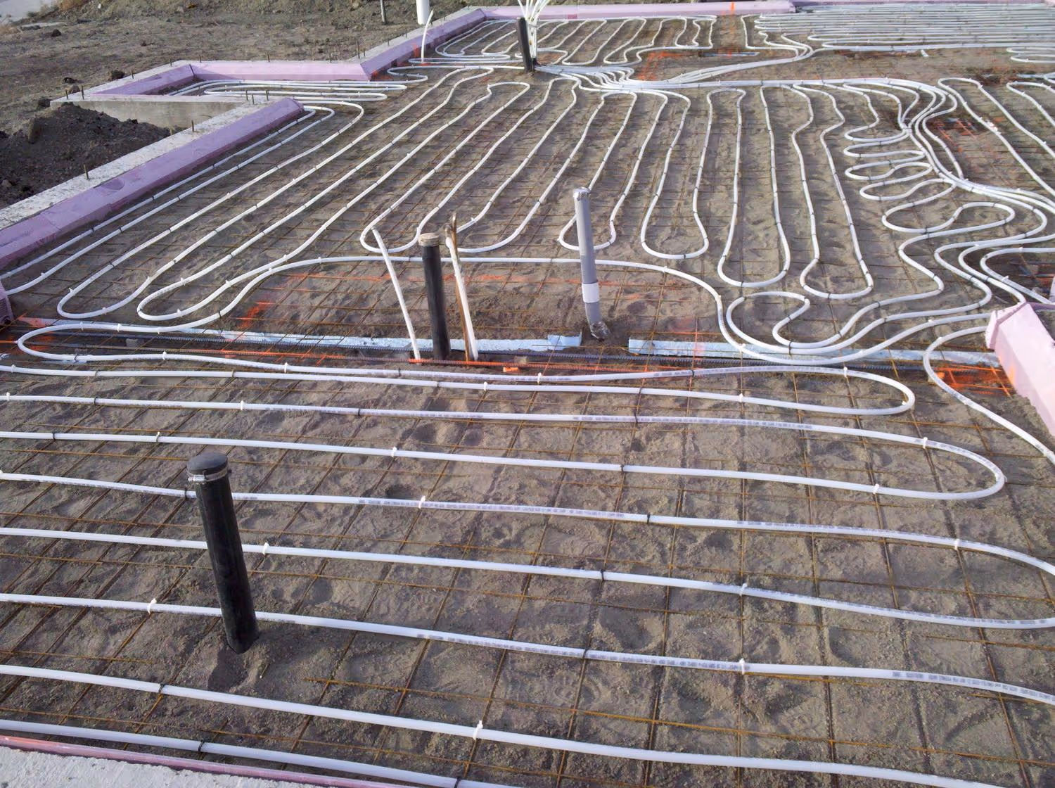 White plastic tubes for underslab radiant heating installed on ground with wire mesh and pink foam insulation edges.