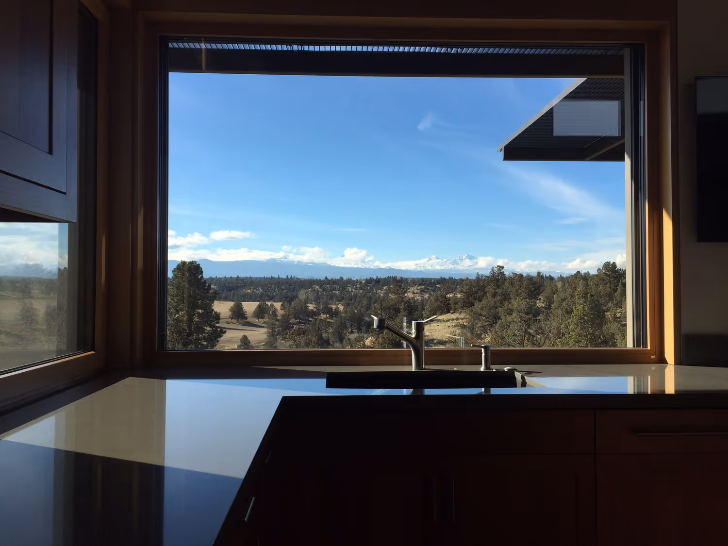 Kitchen sink with window overlooking trees, hills, and snow-capped mountains under a blue sky.