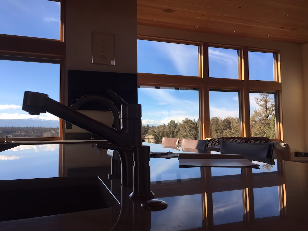 Kitchen faucet silhouette with large windows showing trees and sky reflected on countertop.