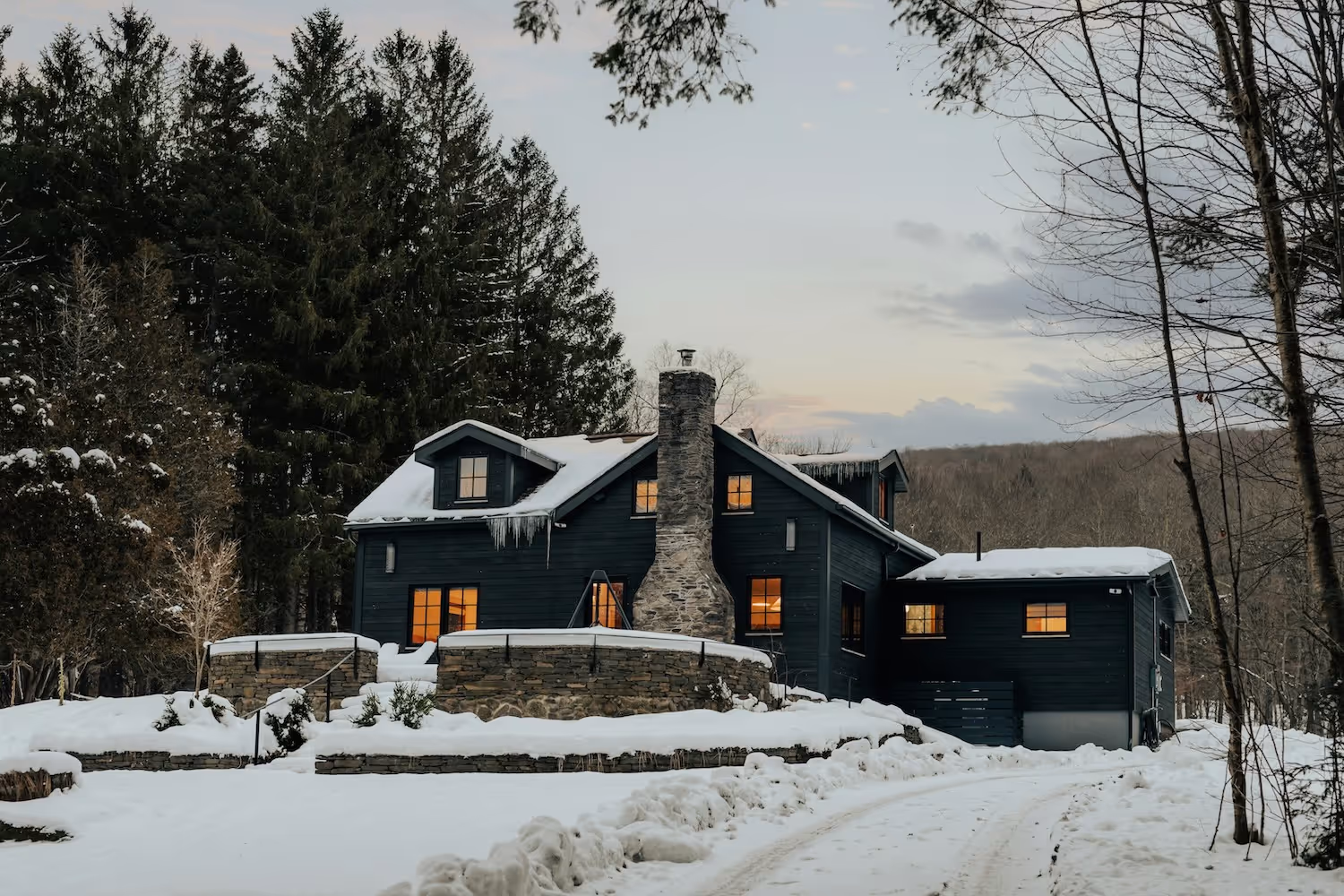 Dark cabin with lit windows surrounded by snow and trees at dusk with snowy driveway in front.