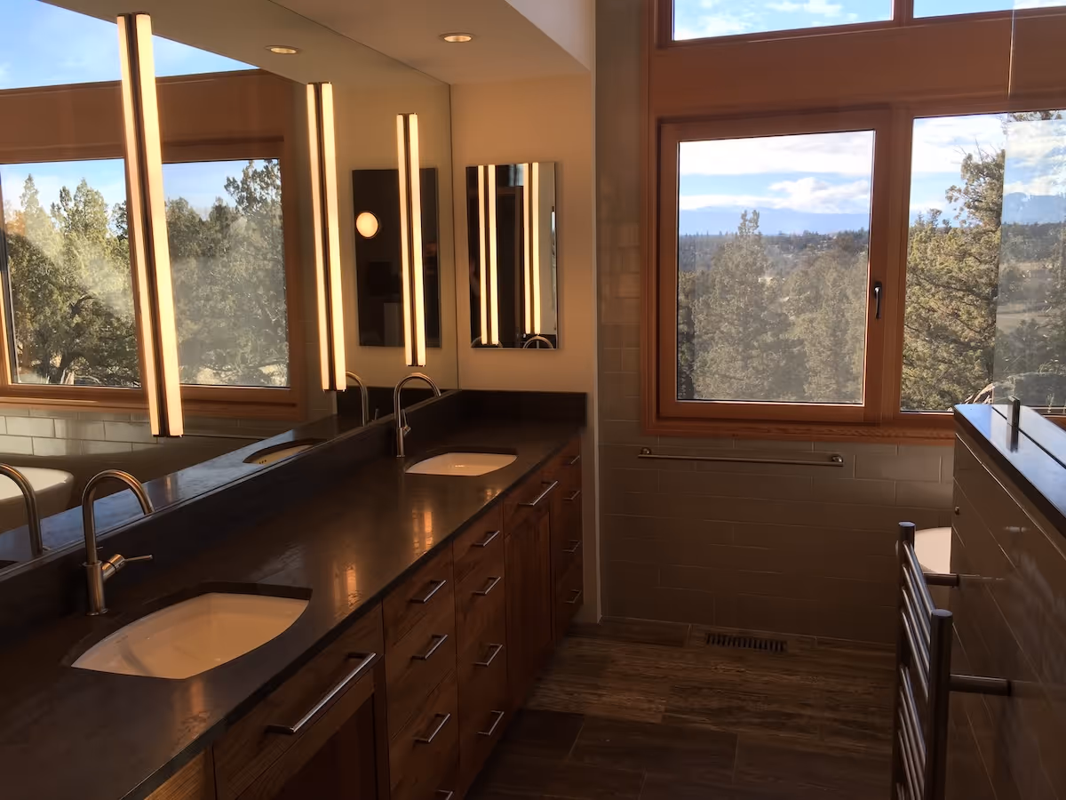 Modern bathroom with double sinks, vertical mirror lights, large window with forest view, and wooden cabinetry.