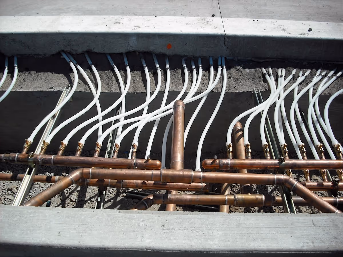 Copper plumbing pipes with multiple white flexible tubes connected beneath a concrete slab.
