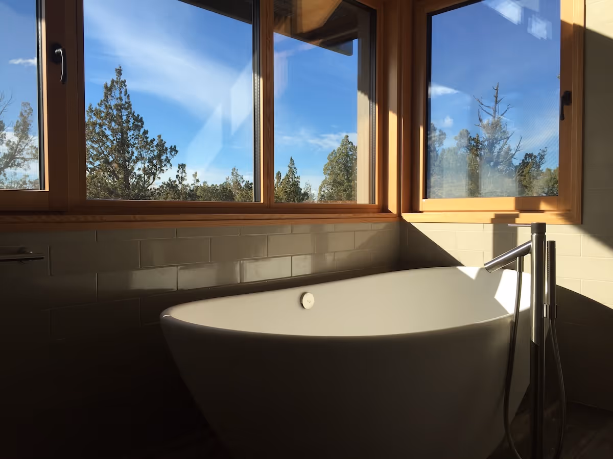 Sunlit modern bathtub near windows showing blue sky and pine trees outside.