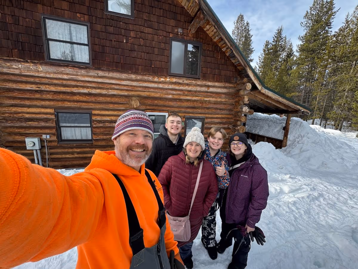 Group of five smiling people wearing winter clothes take selfie outside a snowy log cabin.