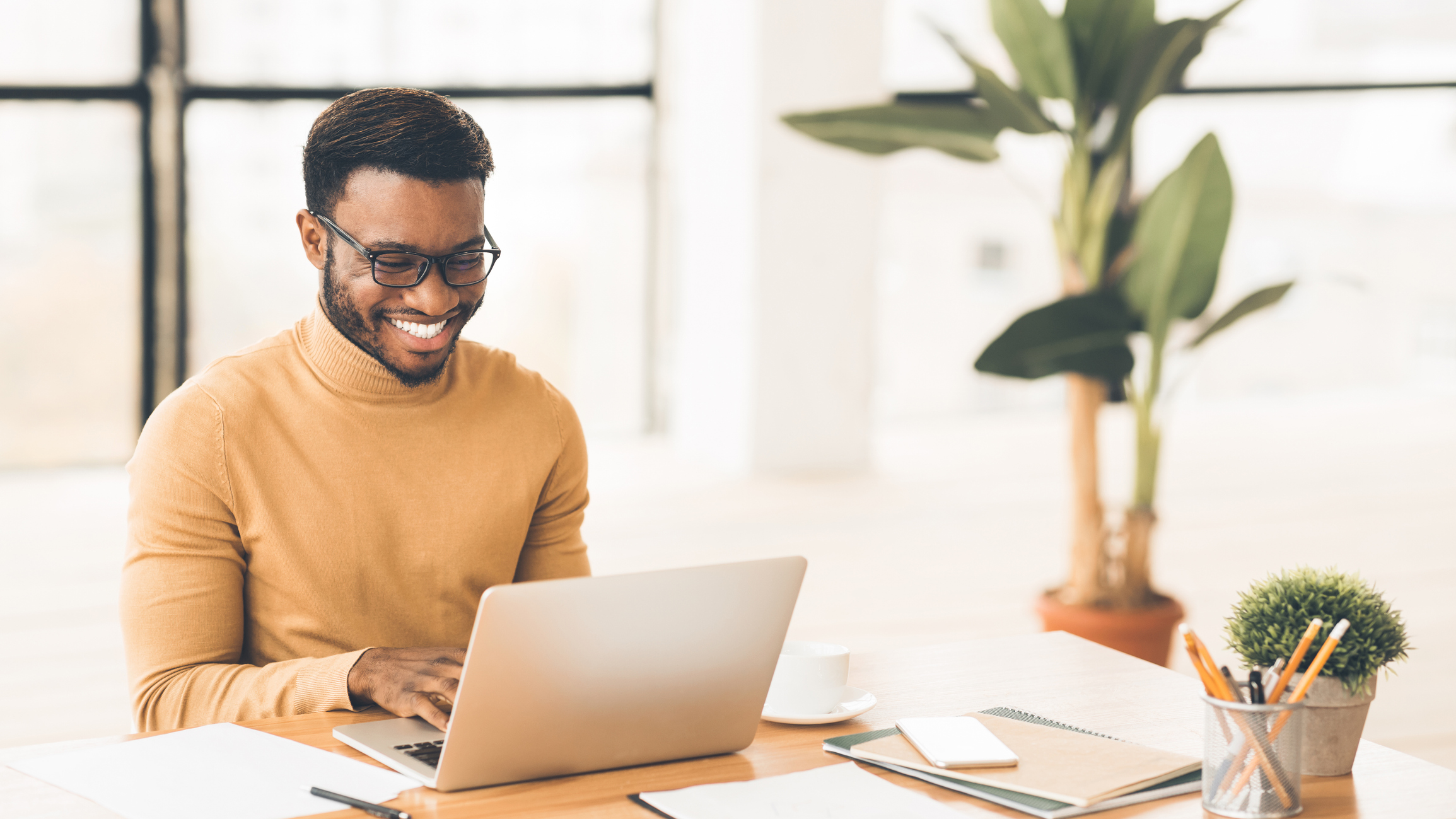 Cheerful man using laptop in a bright office with green plants.