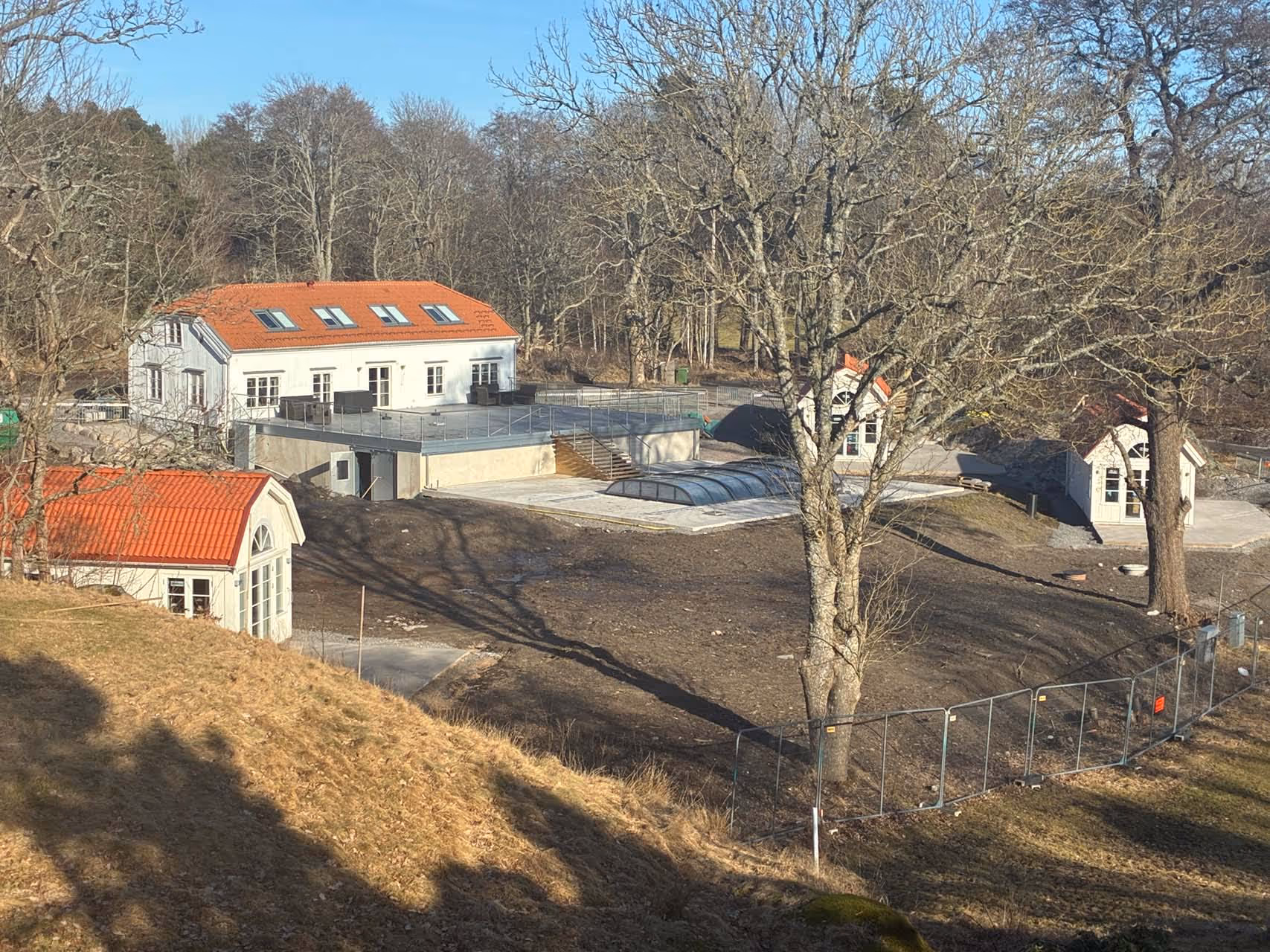 Vit byggnad med röda tegeltak omgiven av lövfria träd och ett inhägnat område under klar himmel.
