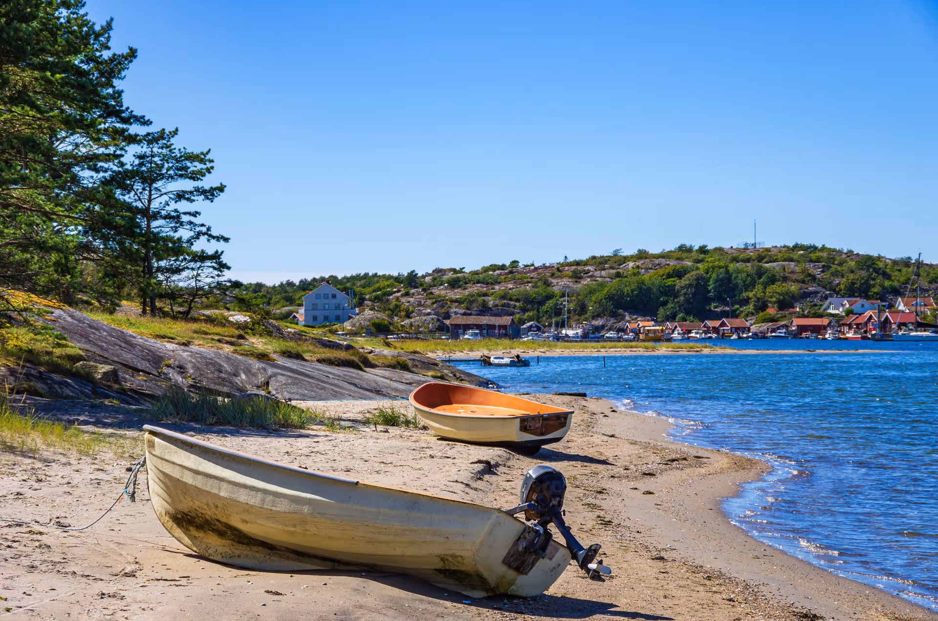 Två små båtar på en sandstrand med klippor, träd och en kust med hus i bakgrunden under en klarblå himmel.