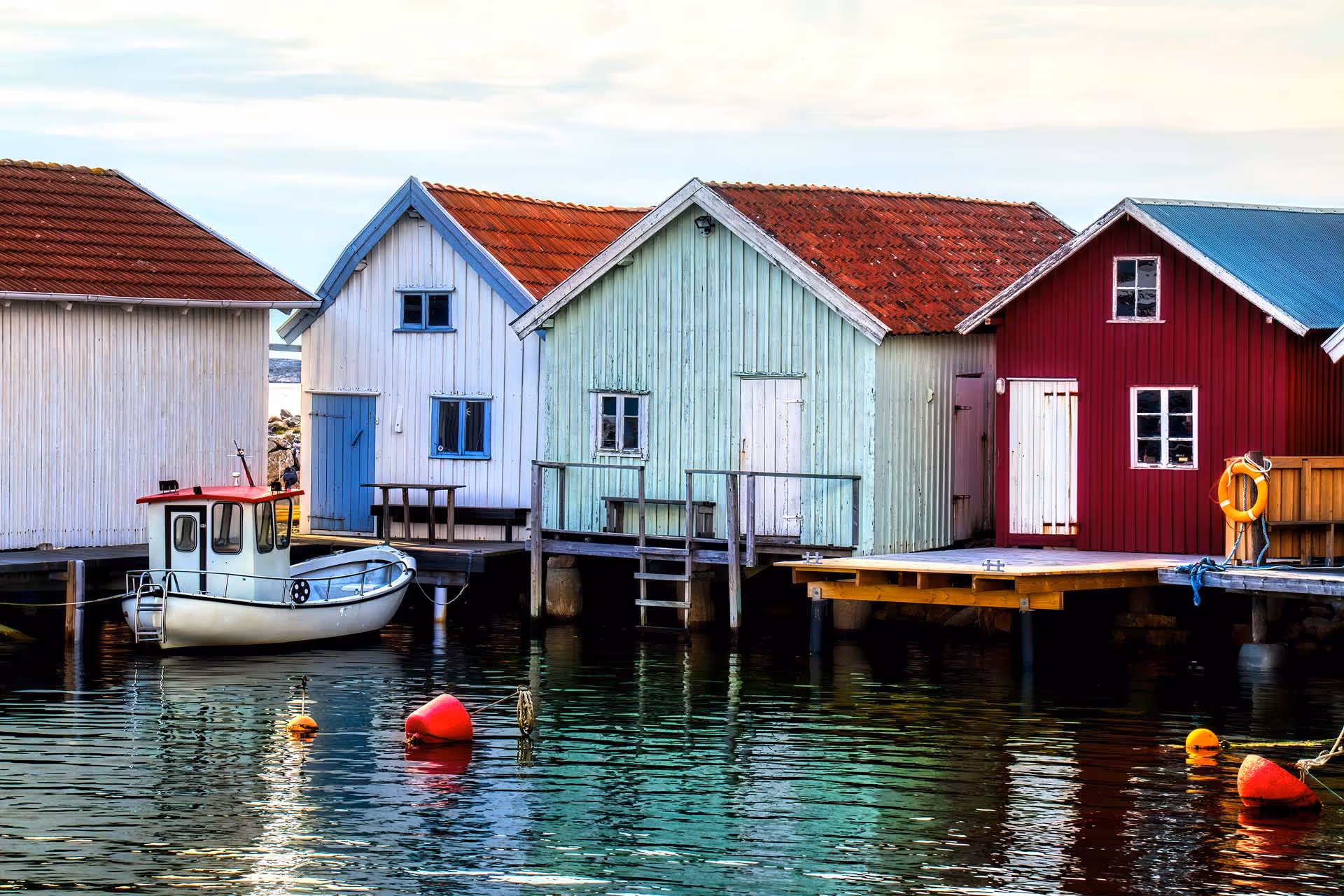Färgglada fiskebodar vid en brygga med en vit båt och röda bojar i vattnet.