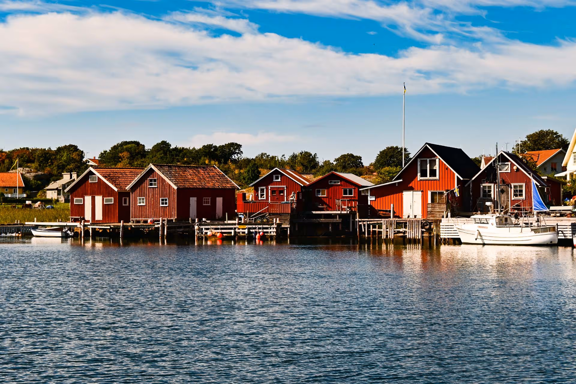 Röda skärgårdshus vid vattnet med en vit segelbåt vid bryggan under en blå himmel med moln.