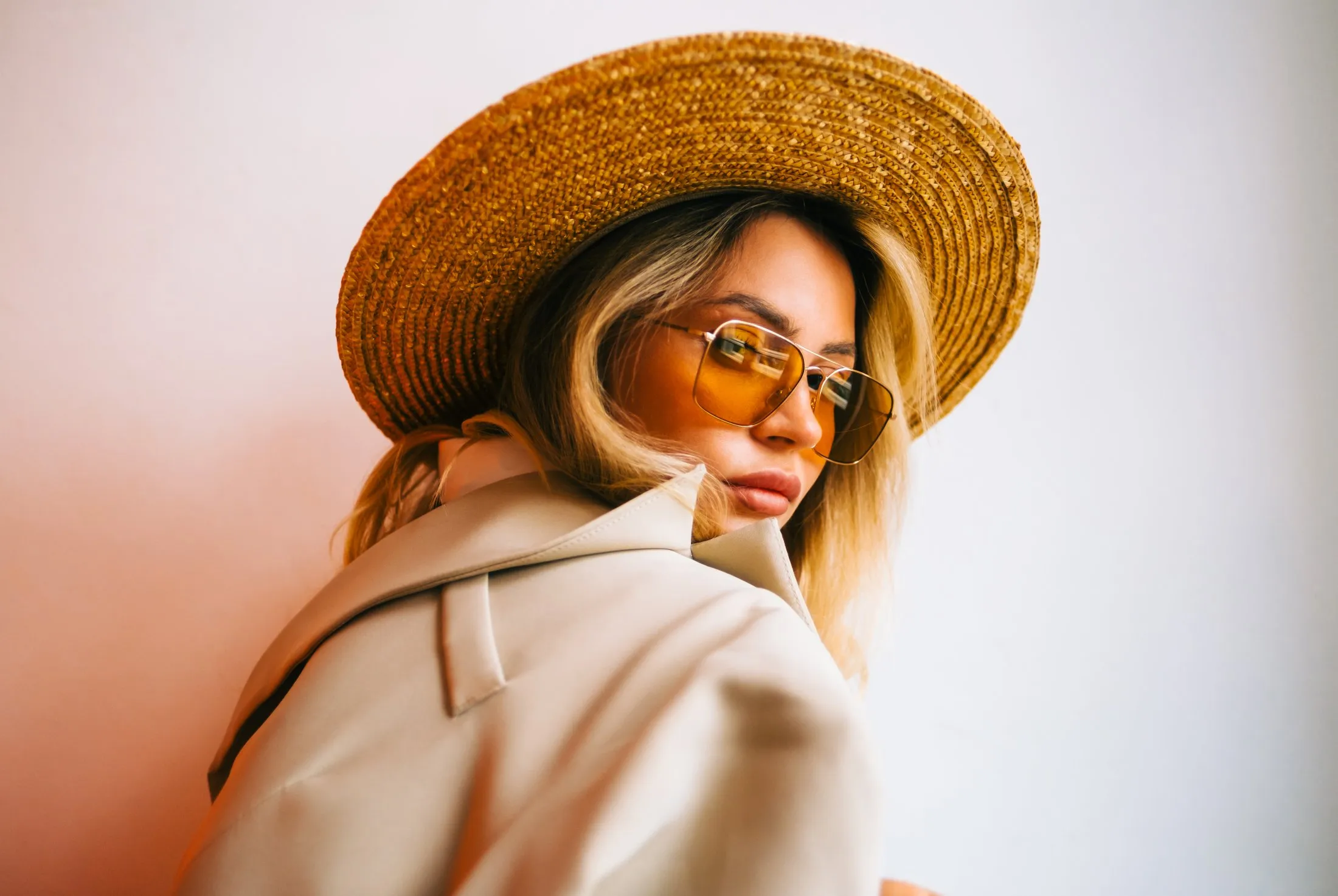 Woman wearing a wide-brimmed straw hat, orange-tinted sunglasses, and a beige coat looking over her shoulder.