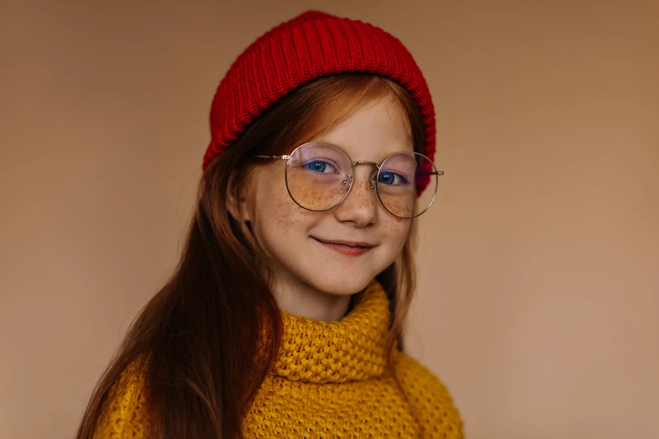 Smiling girl with red hair, freckles, blue eyes, round glasses, red beanie, and yellow knit sweater.