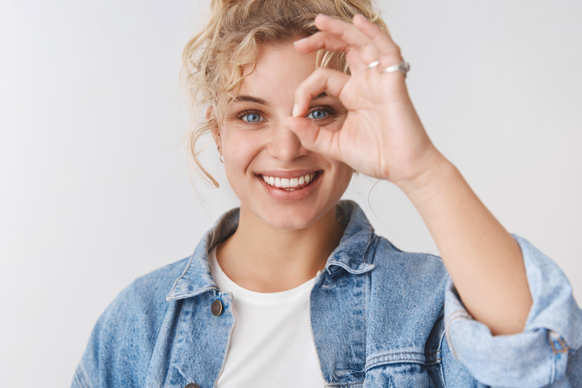 Smiling woman with curly hair looking through an OK hand gesture wearing a denim jacket and white shirt.