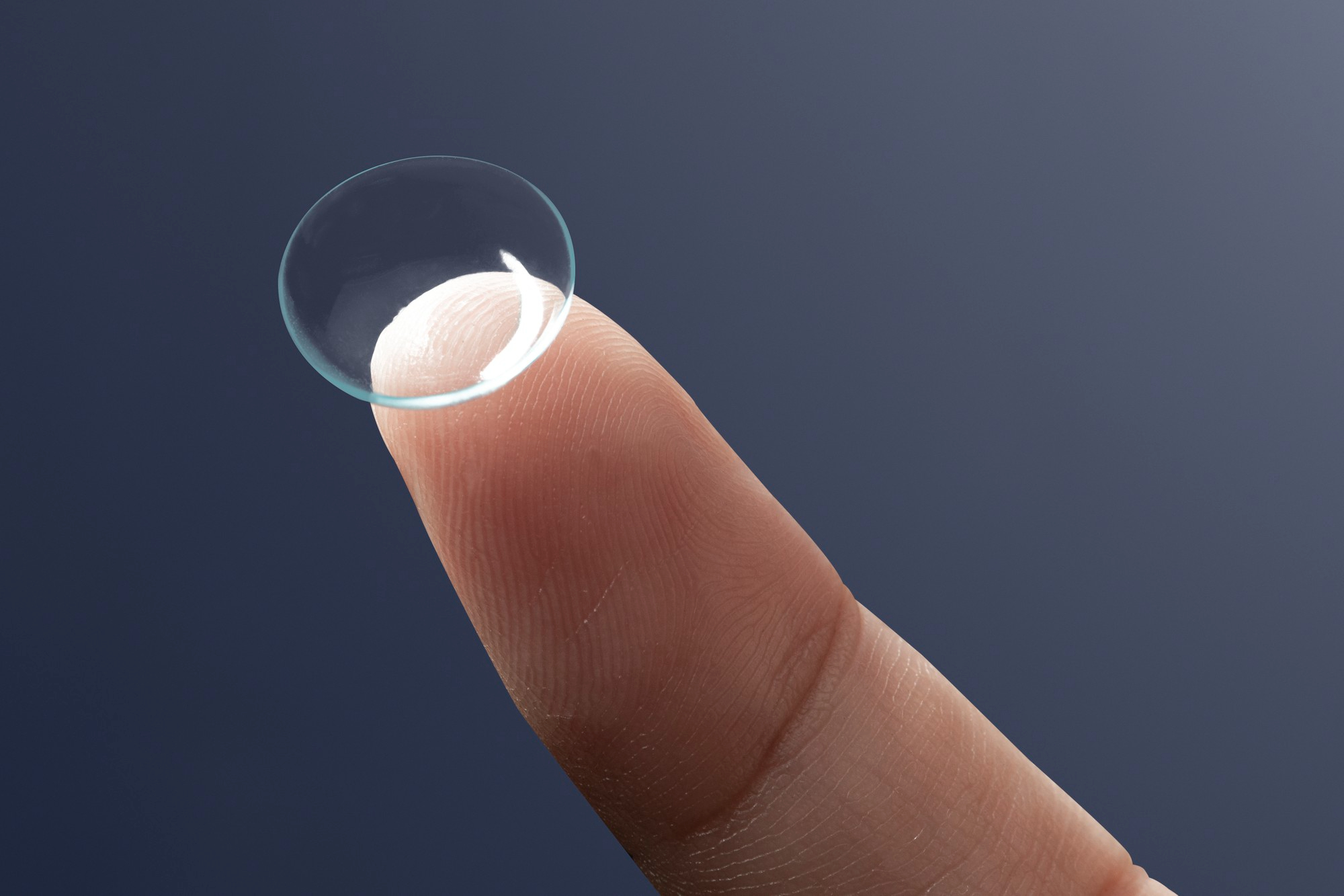 Transparent contact lens resting on the fingertip against a dark background.