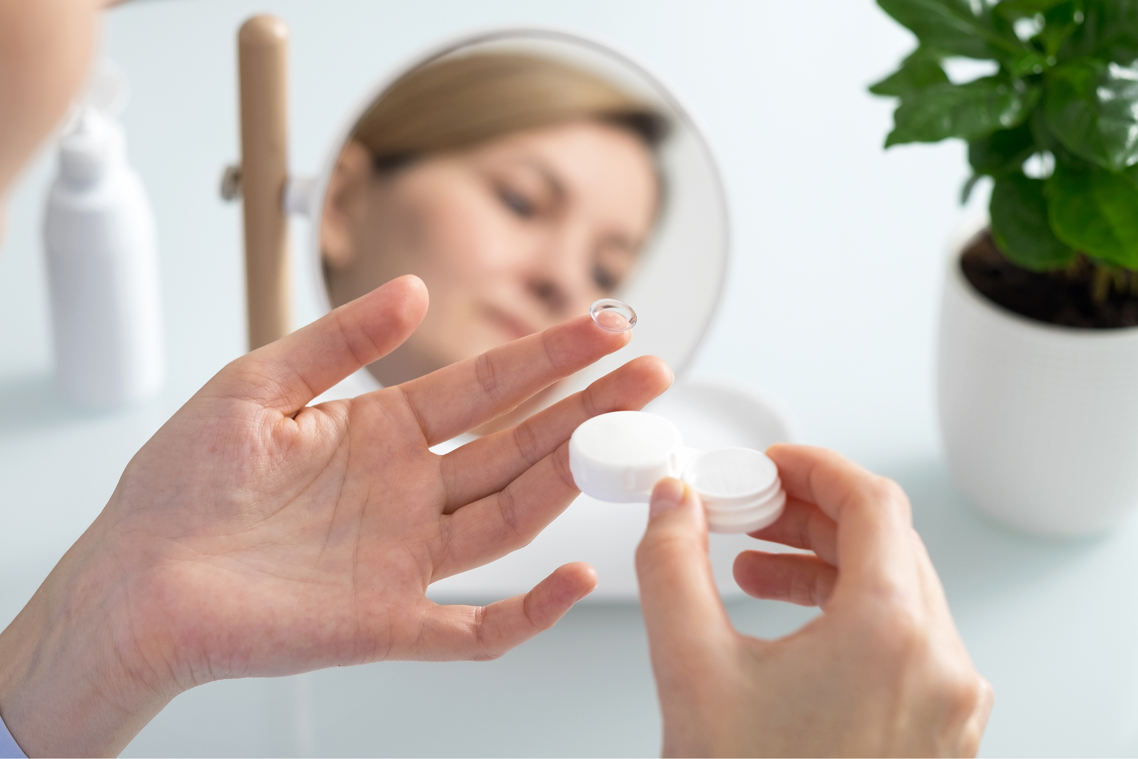 Woman holding a contact lens on her fingertip and a white lens case in her hand with her reflection in a mirror.