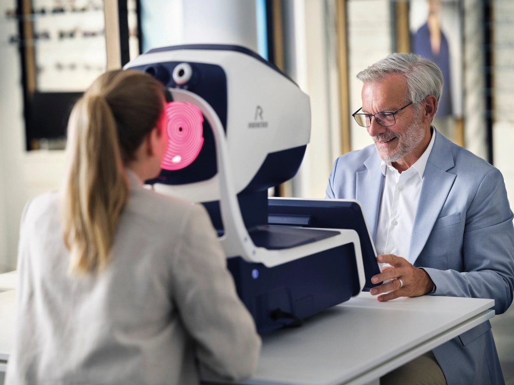 Older man wearing glasses seated at an eye exam machine with a technician guiding the test.