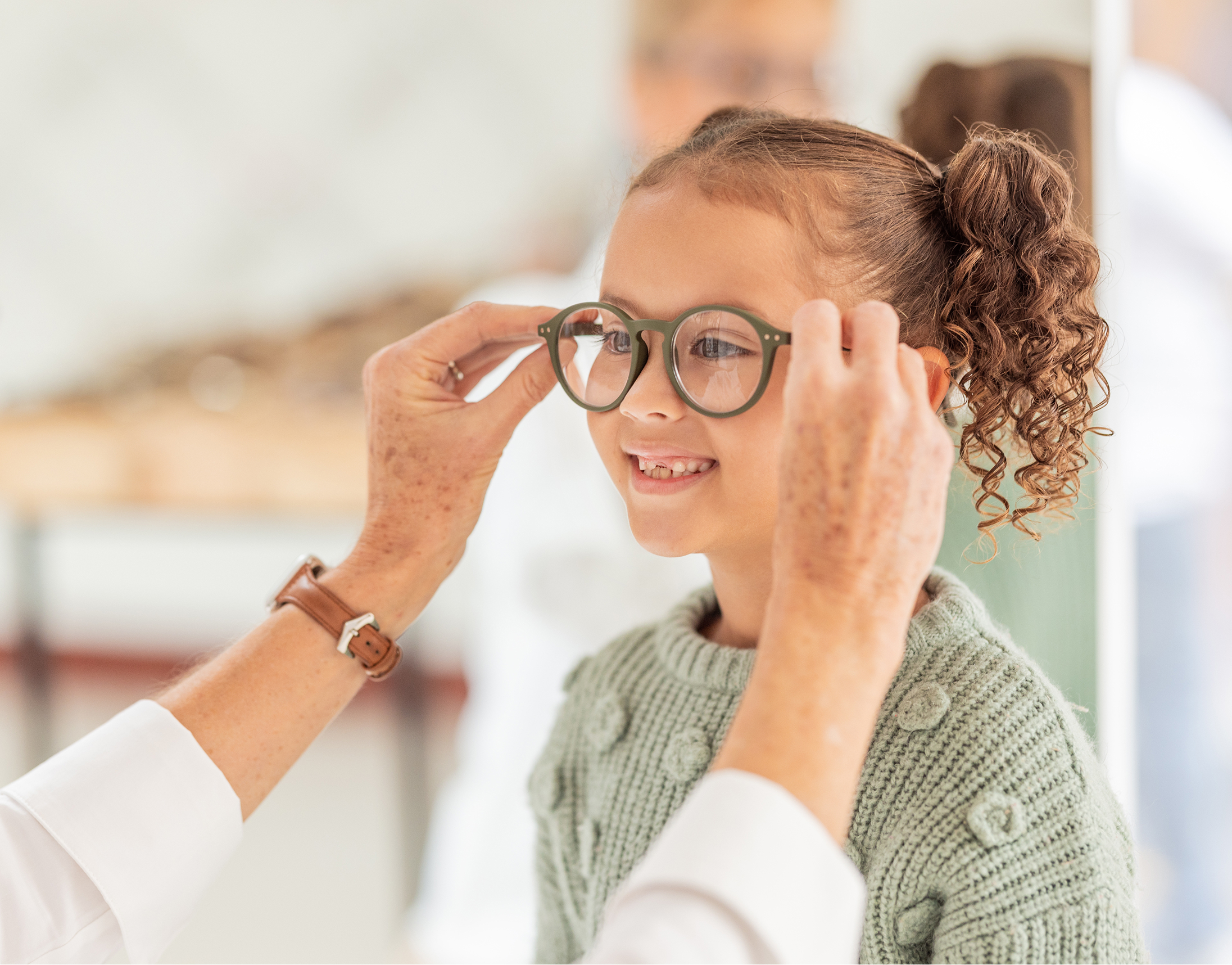 Child smiling while adult adjusts eyeglasses on her face at an eye care appointment.
