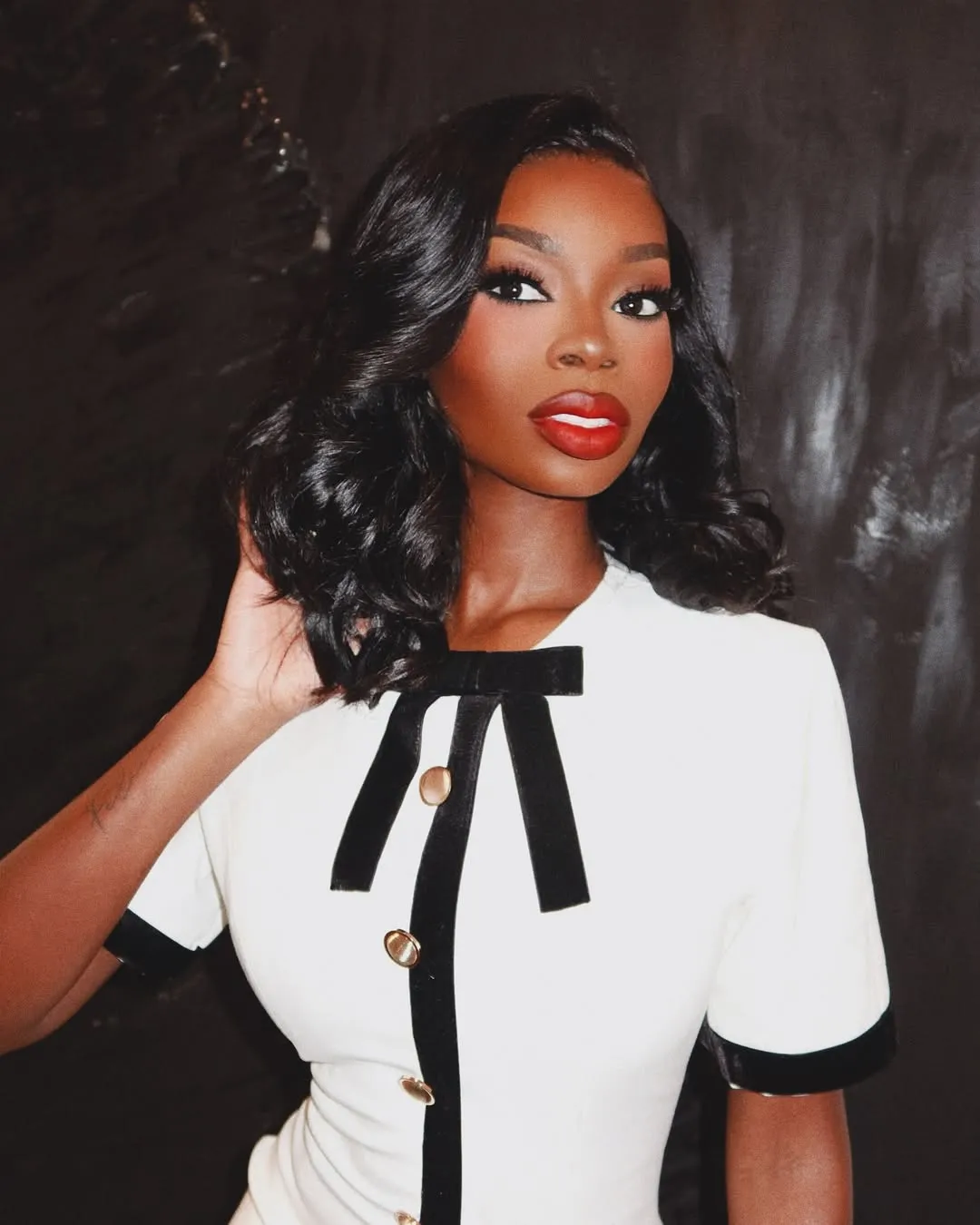 A fitted white mini dress with bold black velvet trim outlining the neckline, centre front, and sleeve cuffs, finished with gold button detail down the front and a delicate black bow at the collar. The structured silhouette and contrast trim give it a distinctly Chanel-coded, ladylike quality — worn here with sheer black tights and black patent pointed-toe heels that sharpen the monochrome palette and extend the leg line cleanly.