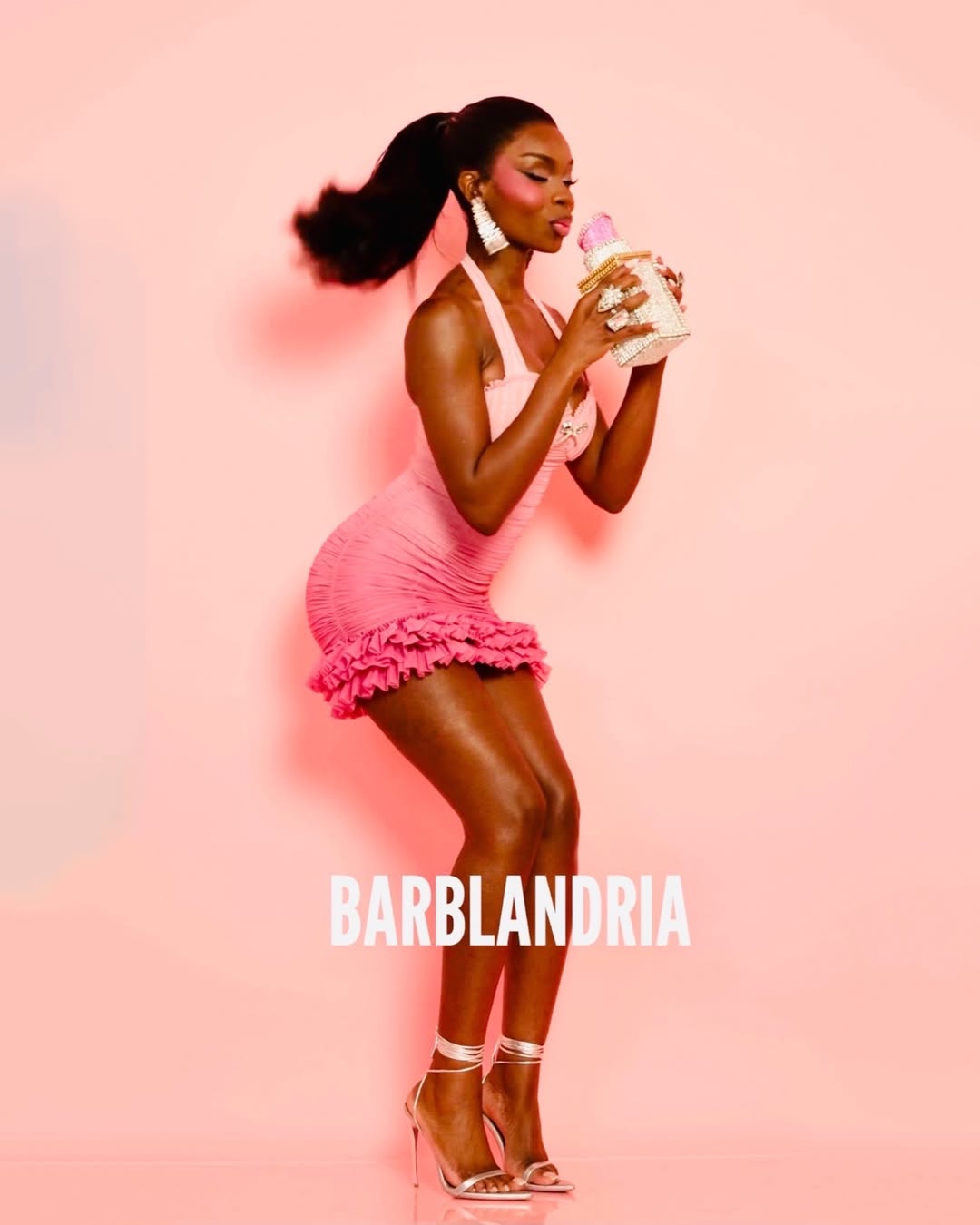 Bubblegum pink halter-neck mini with an all-over ruched body and a tiered ruffle hem, creating a playful, textured silhouette. Accessorised with large white beaded drop earrings, a crystal cocktail ring, silver strappy lace-up stiletto heels, and a novelty cupcake crystal-embellished minaudière clutch with a pink top. High sleek ponytail.