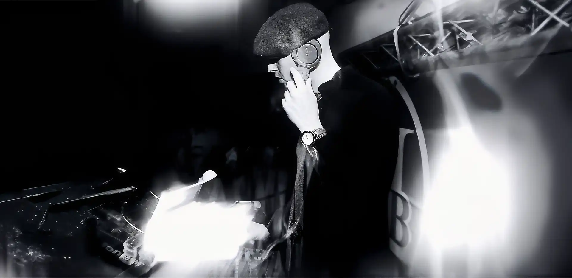 Black and white photo of Neil Pierce DJing wearing a flat cap and headphones, mixing music at a DJ console with bright lights.