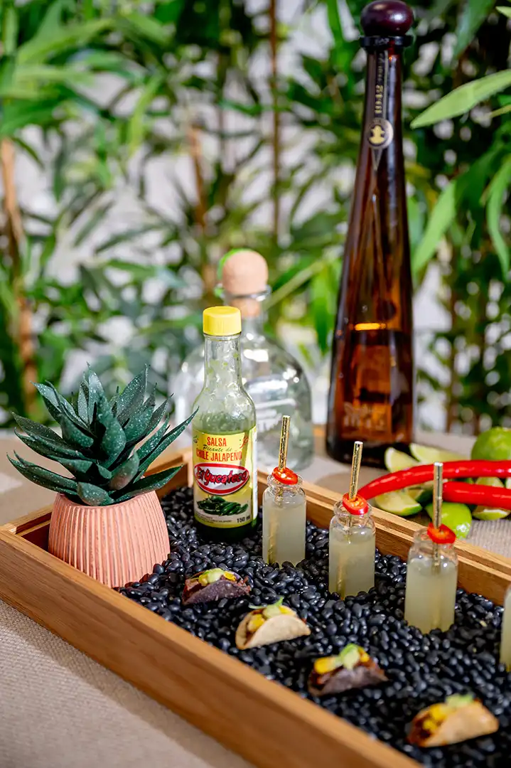 Wooden tray filled with black beans holding mini tacos, three small shot glasses with straws and chili slices, a bottle of green jalapeño hot sauce, a small potted succulent, and tequila bottles in the background.