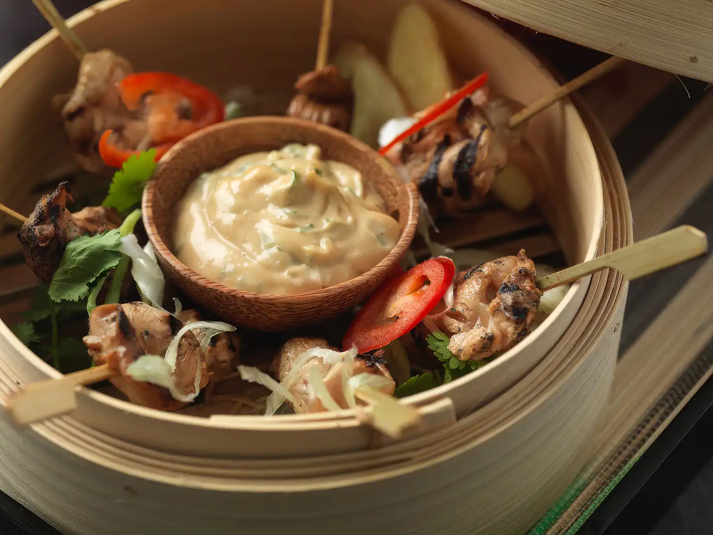 Grilled chicken skewers with sliced red chili and herbs around a bowl of creamy dipping sauce in a bamboo steamer basket.