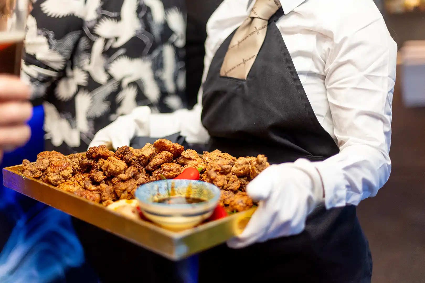 Waiter in white gloves and black apron holding a tray of fried chicken pieces with a small bowl of dipping sauce.
