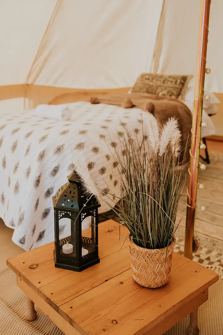 Wooden table with a woven basket containing decorative grass and a black lantern with two candles, next to a bed with patterned bedding and pillows.