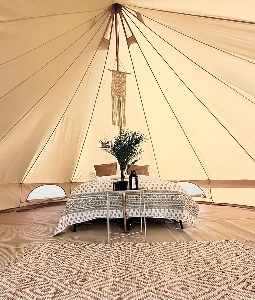 Interior of a beige bell tent with a bed, patterned bedspread, two brown pillows, a small table holding a potted palm and a lantern, with a woven rug in the foreground.