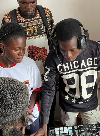 Four young people, from developing countries, gathered around DJ equipment, one wearing headphones and a black Chicago 98 shirt operating the controls.