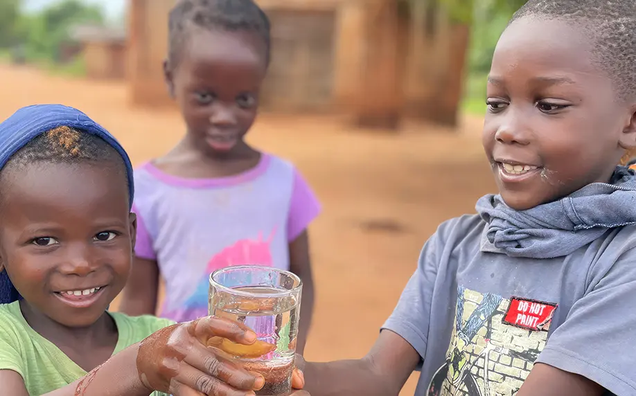 Two smiling children holding a glass of water together with a third child in the background.
