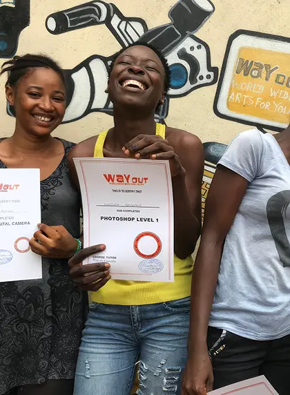 Three smiling young women holding WayOut certificates, one showing a Photoshop Level 1 certificate, standing in front of a mural.