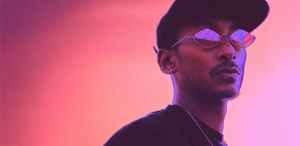 Jitwam wearing a dark coloured cap and geometric sunglasses with a reflective Gucci pattern, against a pink and orange gradient background.