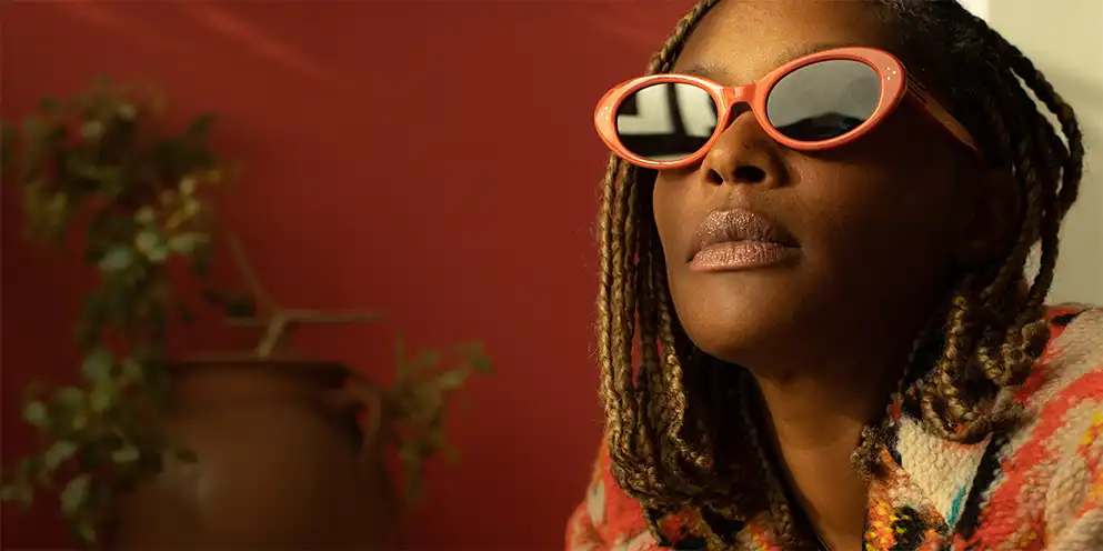 Megatronic with her braided hair wearing coral sunglasses and a patterned fleece jacket indoors against a red and white wall.