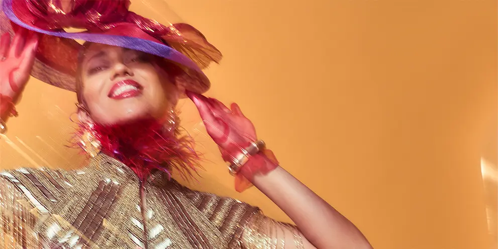 Natasha Diggs wearing a purple hat, red gloves, and a fringed gold outfit smiling against an orange background.